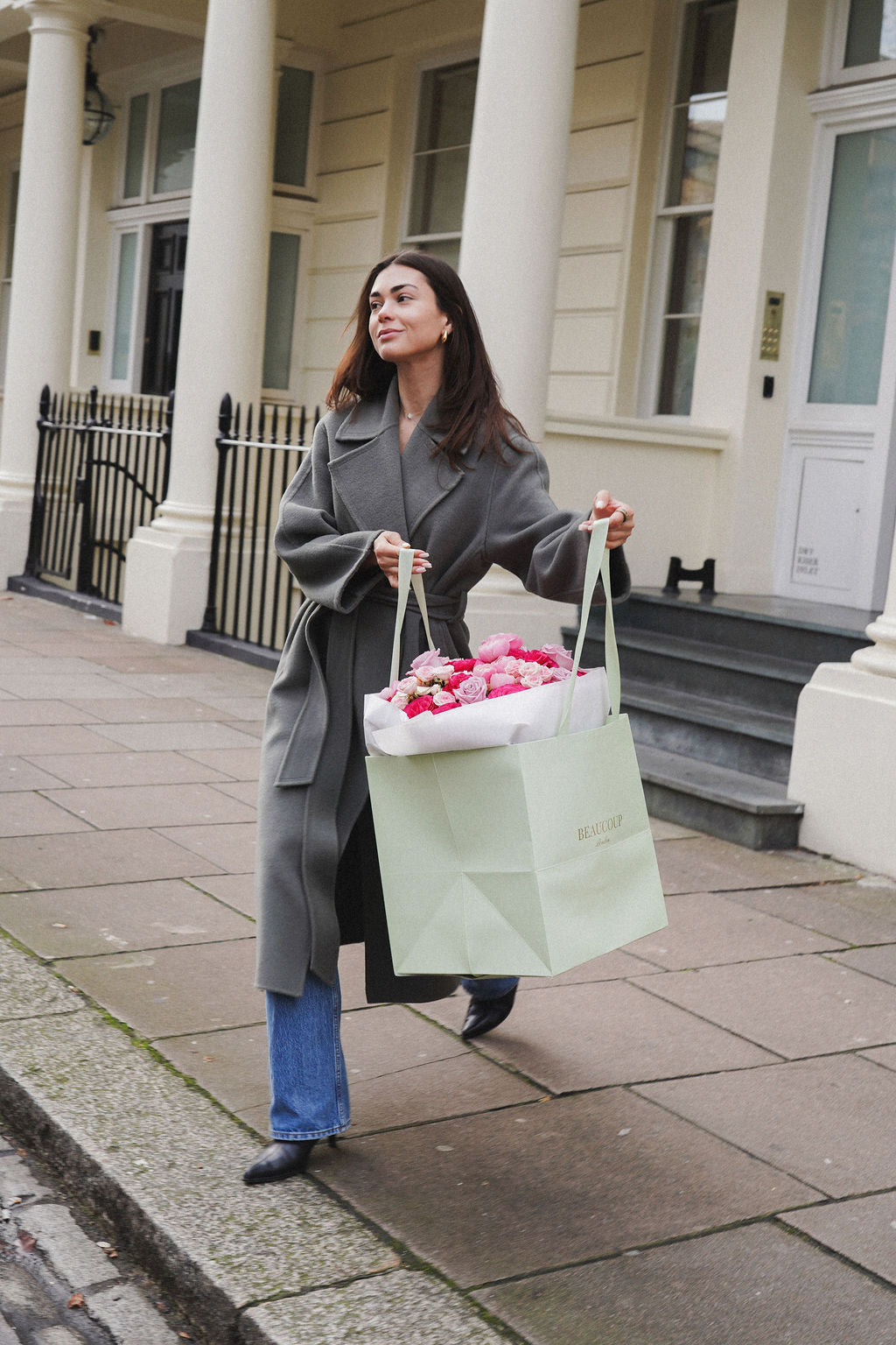 Woman in a gray coat holding a light green bag with flowers, standing on a sidewalk.